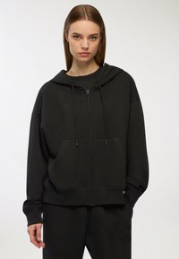 Czarna bluza z kapturem zapinana na zamek, z kieszeniami z przodu i sznurkiem. Miękka tkanina, luźny krój, ribbowe mankiety i dół. Prosty, jednolity design.