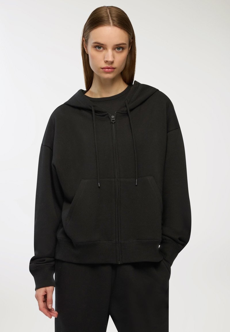 Czarna bluza z kapturem zapinana na zamek, z kieszeniami z przodu i sznurkiem. Miękka tkanina, luźny krój, ribbowe mankiety i dół. Prosty, jednolity design.