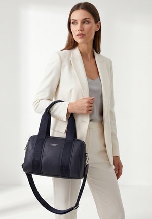 Femme en costume crème tenant un sac à main en cuir noir texturé avec une bandoulière et des logos de marque visibles dans un cadre minimaliste.