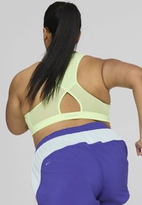 Svetlozeleni športni nedrček s križnim hrbtom in mrežastimi paneli. V kompletu s vijoličnimi kratkimi hlačami z belim elastikom.