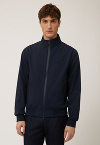Giacca leggera blu navy con alto colletto, zip completa e polsi a costine. Due tasche laterali e dettaglio del logo discreto sul petto.