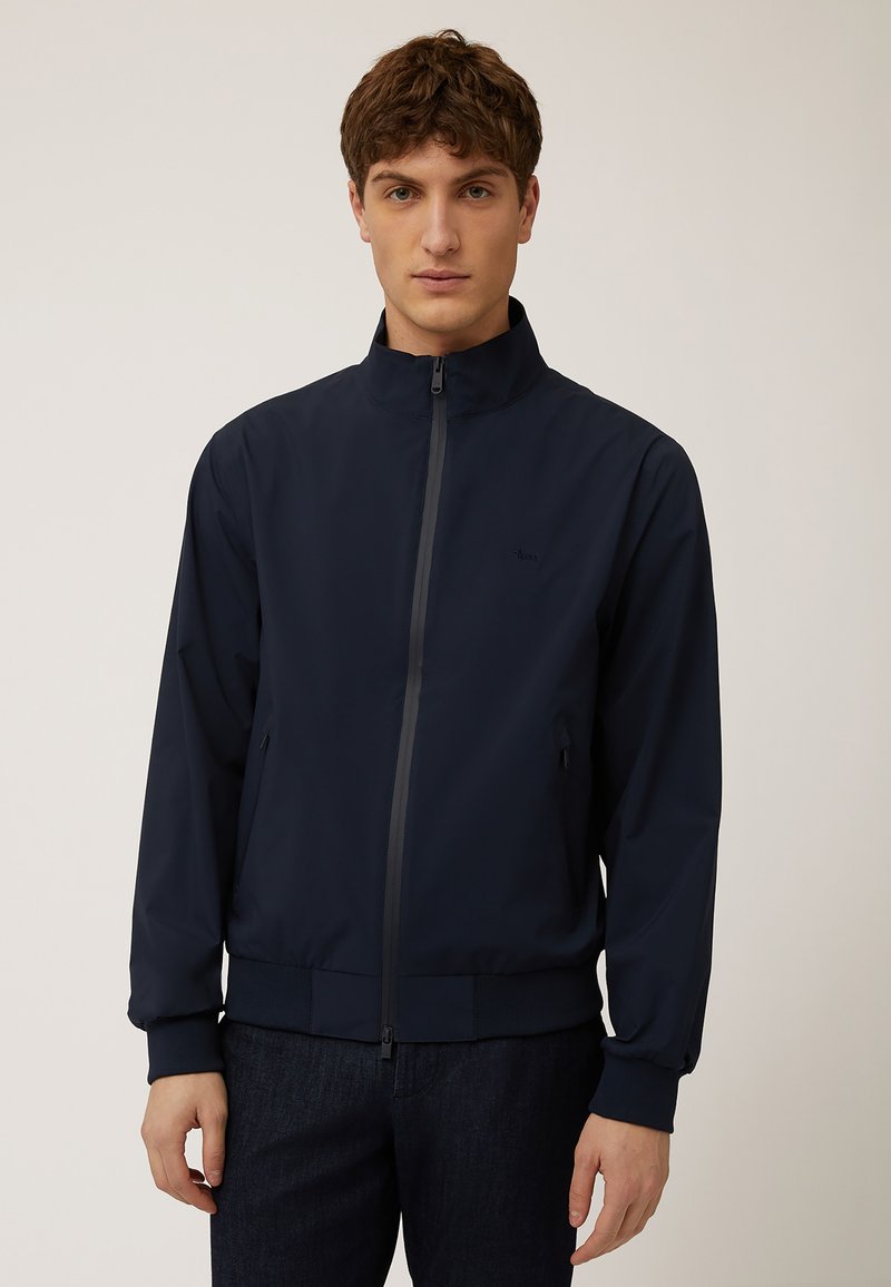 Giacca leggera blu navy con alto colletto, zip completa e polsi a costine. Due tasche laterali e dettaglio del logo discreto sul petto.
