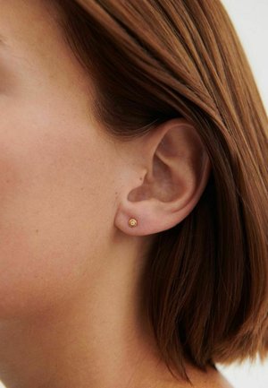 Close-up of a woman's ear with short brown hair wearing a small round gold stud earring with a clear gemstone.