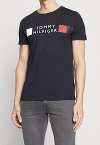 Man wearing black Tommy Hilfiger t-shirt with white and red logo and gray jeans, standing with arms relaxed by sides.