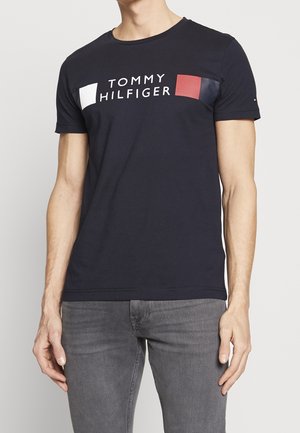 Mężczyzna w czarnym t-shircie Tommy Hilfiger z białym i czerwonym logo oraz szarych dżinsach, stojący z rękami swobodnie wzdłuż ciała.
