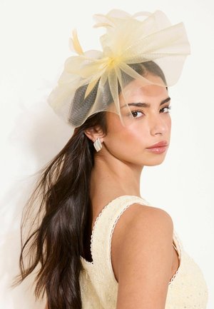 Woman with long dark hair wearing a cream textured sleeveless dress and a large pale yellow mesh fascinator with feather details.