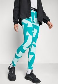 Leggings moulants en teal avec des motifs abstraits de flèches bleu clair, une taille élastique noire, associés à des chaussures de sport noires.