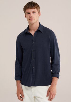 Jeune homme portant une chemise bleu marine à boutons avec les manches retroussées et un pantalon blanc, debout devant un fond beige uni.