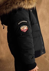 Melna pūkainā jaka ar kažoku apādē. Tajā ir auduma uzplecs ar "SUPERDRY®" un "EXPEDITION EVEREST" uz kreisās piedurknes. Tekstūrēti aproces.