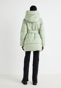 Manteau molletonné vert menthe avec capuche, taille ceinturée et texture matelassée. Présente une coupe mi-longue et des manches longues. Porté sur un pantalon sombre.