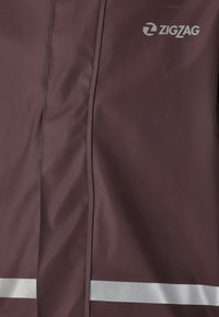 Chaqueta impermeable burdeos con una textura suave, con franjas reflectantes plateadas y un logo en zigzag en la parte superior izquierda.