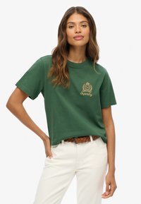 T-shirt en coton vert à manches courtes, avec un logo en broderie dorée sur la poitrine, associé à un pantalon de couleur crème et une ceinture tressée.
