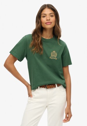 T-shirt en coton vert à manches courtes, avec un logo en broderie dorée sur la poitrine, associé à un pantalon de couleur crème et une ceinture tressée.