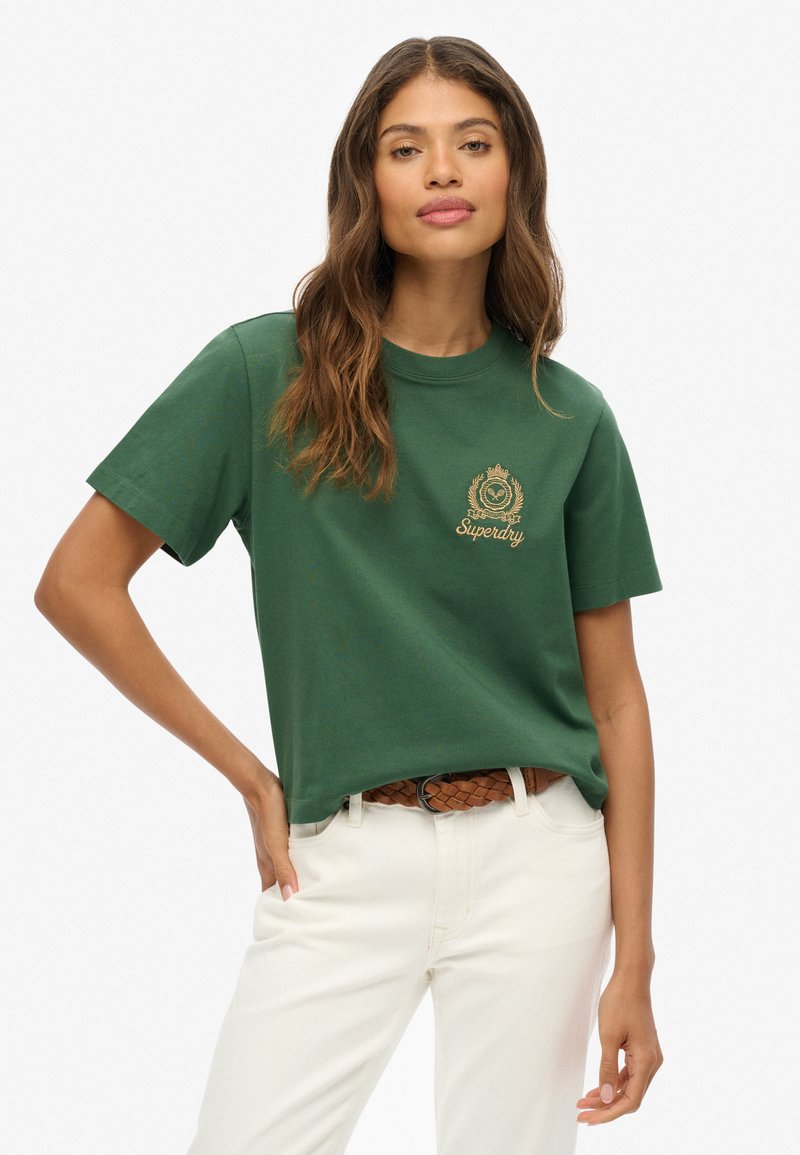 T-shirt en coton vert à manches courtes, avec un logo en broderie dorée sur la poitrine, associé à un pantalon de couleur crème et une ceinture tressée.