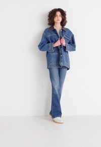 Denim jacket with large pockets, a brown collar, and blue flared jeans. Worn with light sneakers, set against a plain white background.