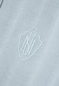 Un tissu tricoté bleu clair présente un tissage texturé avec un logo écusson brodé. Le design est minimaliste et uniforme en couleur.