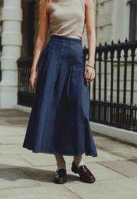 Tamsiai mėlynas džinsinis maksi sijonas su klostėmis, derinamas su tamsiomis loafer batais ir šviesiomis kojinėmis, fone matoma urbanistinė aplinka.