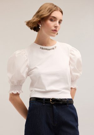 T-shirt bianco con maniche a sbuffo e polsini arricciati, impreziosito da perline metalliche al collo, abbinato a jeans scuri a vita alta e una cintura nera.
