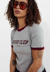 Pelēks t-krekls ar bordo krāsas apkakli un piedurkņu akcentiem, uz kura ir uzraksts "GOSSIP CLUB" un dibināšanas gads 1992 tumši bordo krāsā.