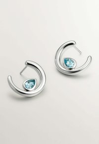 Pendientes de aro de plata con un diseño escultórico, que presentan una gema en forma de lágrima de color azul claro, engastada en un marco de metal pulido.