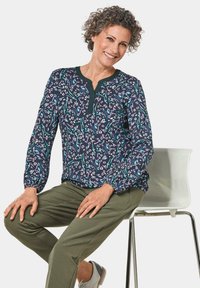 Marinebluse mit floralem Muster in Grün und Weiß, runder Ausschnitt mit Knopfleiste, lange Ärmel mit gerafften Bündchen, kombiniert mit grünen Hosen.