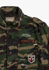 Giacca militare mimetica in tonalità di verde e marrone con colletto pieghevole, patch "U.S.A. ARMY" e distintivo "THUNDER FLIGHT" sulla tasca.