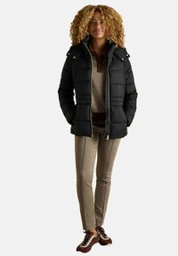 Must mustjakk kapuutsiga, tikitud disainiga, kuldsete detailidega, beež mustriga ülisärki, pruunide ruuduliste pükste ja pruunide spordijalatsitega.