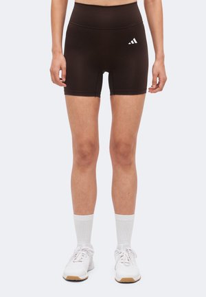 Femeie purtând pantaloni scurți sport negri cu talie înaltă și logo alb, șosete albe și pantofi de alergare albi, stând drept pe fundal deschis.