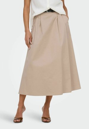 Žena oblečená v béžovej midi sukni áčkového strihu so záhybmi, čiernym opaskom, bielou blúzkou a hnedými sandálmi s otvorenou špičkou, stojaca so založenými rukami v vreckách.