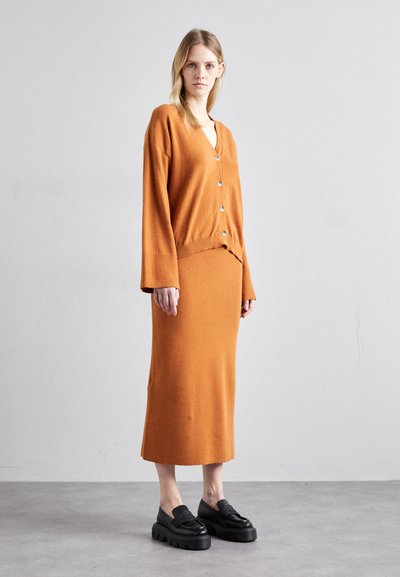 Ensemble en deux pièces en tricot orange, comprenant un long cardigan avec des boutons et une jupe midi assortie. Associé à des chaussures noires massives.