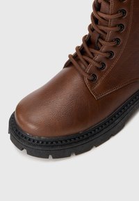 Brun fusk-läderankelboot med rund tå, svart gummisula och svarta metallöglor. Har texturerat material och snörningsdesign.