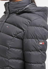 Černá péřová bunda s vysokým límcem, s prošívanými sekcemi a bočním logem. Materiál vypadá hladce a leskle.