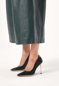 Fekete lakkbőr magas sarkú cipő hegyes orral, karcsú, arany díszítésű tűsarkúval, zöld bőrszoknyával párosítva.