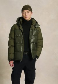 Πράσινο μπουφάν puffer με κουκούλα, κατασκευασμένο από γυαλιστερό υλικό και με σχέδιο καπιτονέ. Φορεμένο πάνω από μαύρο ζιβάγκο και σε συνδυασμό με σκούρο παντελόνι cargo.