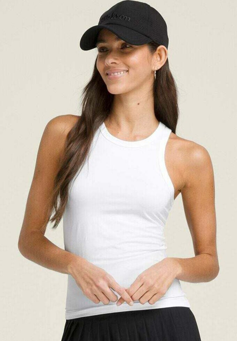 Frau mit langem braunem Haar, die eine schwarze Mütze, ein weißes ärmelloses Oberteil und einen schwarzen Rock trägt, lächelt und hält die Hände vor sich verschränkt.