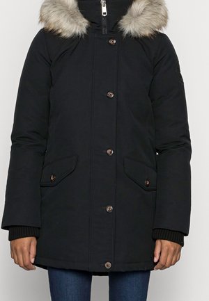 Manteau d’hiver noir avec fermeture à boutons et à glissière, poches à rabat et capuche bordée de fourrure, porté sur un jean bleu.