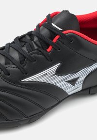 Scarpa sportiva nera sintetica con accenti rossi e lacci neri. Presenta un lato testurizzato con un design del logo a strisce bianche.
