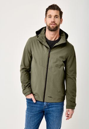 Man with beard wearing olive green zip-up jacket and blue jeans, standing with one hand in pocket against plain background.