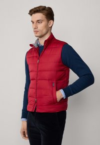 Chaleco acolchado rojo con cierre de cremallera, cuello alto y forro gris. Presenta bolsillos laterales y un corte slim, usado sobre una camiseta de manga larga azul marino.