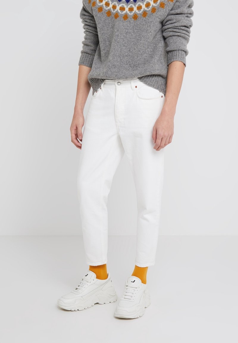 Vita denimjeans med rak passform, kombinerat med en grå stickad tröja med en färgglad cirkulär design och vita sneakers med orangea strumpor.