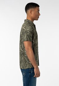 Kurzarm-Hemd in Olivgrün mit einem Blattmuster, ausgestattet mit einem Knopfkragen und einem geraden Saum. Getragen mit blauen, ausgewaschenen Jeans.