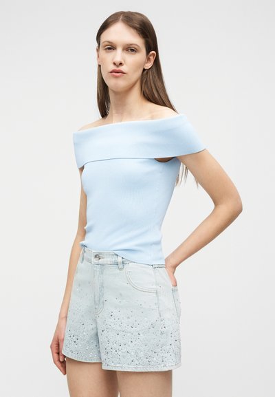 Νεαρή γυναίκα με ανοιχτό μπλε τοπ χωρίς ώμους και ανοιχτόχρωμο σορτς από τζιν με παγιέτες, στέκεται με το ένα χέρι στη μέση της.