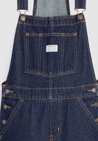 Salopette en jean bleu foncé avec poche poitrine avant, bretelles réglables avec boutons métalliques et fermetures latérales à boutons.