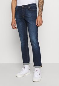 Jeans de mezclilla azul oscuro con corte ajustado, que presentan un diseño clásico de cinco bolsillos, puños doblados y una pretina cosida.