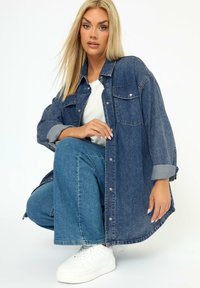 Camicia di jeans blu scuro, vestibilità ampia, con chiusura a bottoni e tasche sul petto. Abbinata a pantaloni in denim blu chiaro e scarpe da ginnastica bianche.