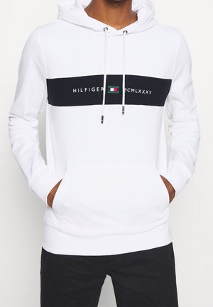 Osoba nosząca białą bluzę z kapturem z czarnym poziomym paskiem i logo Tommy Hilfiger, ręce w przedniej kieszeni, w połączeniu z czarnymi spodniami.