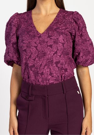 Femme portant un chemisier violet profond à motif floral texturé et manches courtes bouffantes, associé à un pantalon taille haute cintré assorti.