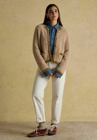 Bež villase cardigan kuldnuppudega, denimipusa all, valgete pükste ja mitmevärviliste tossudega, reljeefse taustaga.