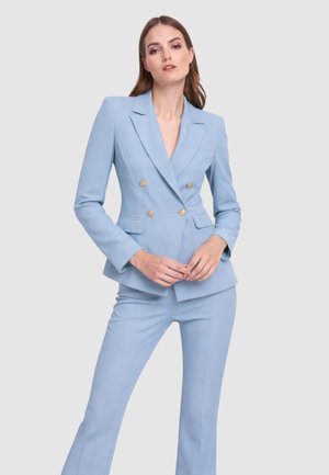 Donna che indossa un blazer doppiopetto azzurro chiaro con bottoni dorati e pantaloni su misura abbinati, in piedi davanti a uno sfondo semplice.