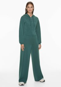 Sweat à capuche zippé vert avec poches avant, associé à un pantalon à jambes larges assorti. Texture lisse, design décontracté. Accentué par des cordons de serrage et des chaussures contrastées.